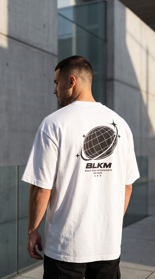 A man viewed from behind wearing a white t-shirt with a black graphic of a globe and the text 'BLKM' on the back, standing outdoors in front of a modern concrete building.