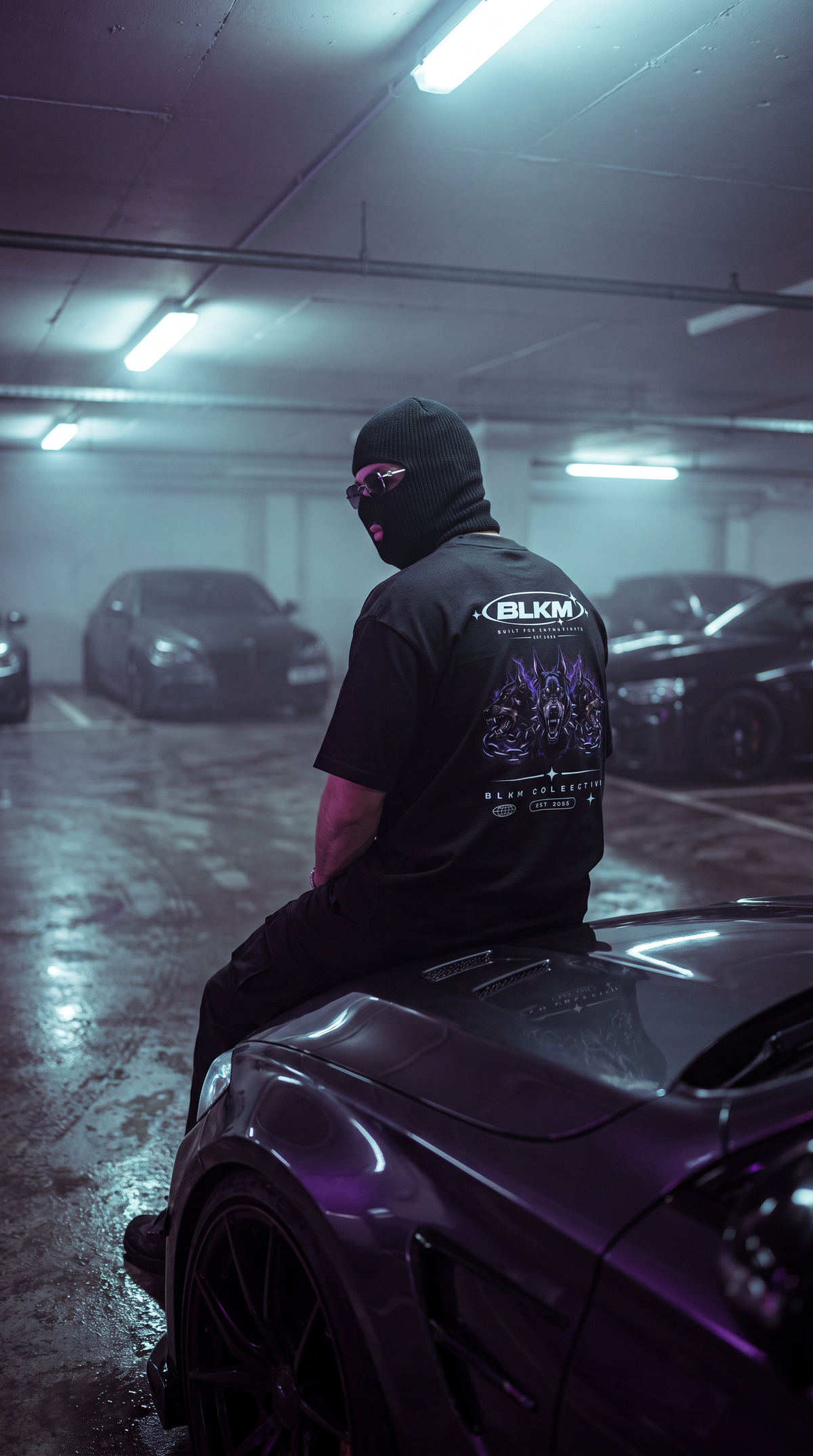A person wearing a black hoodie with a face mask and sunglasses sits on the front fender of a dark purple sports car in a misty, dimly lit parking garage. The back of the black t-shirt features a graphic print with the brand name 'BLKM' visible on the back of the hoodie.