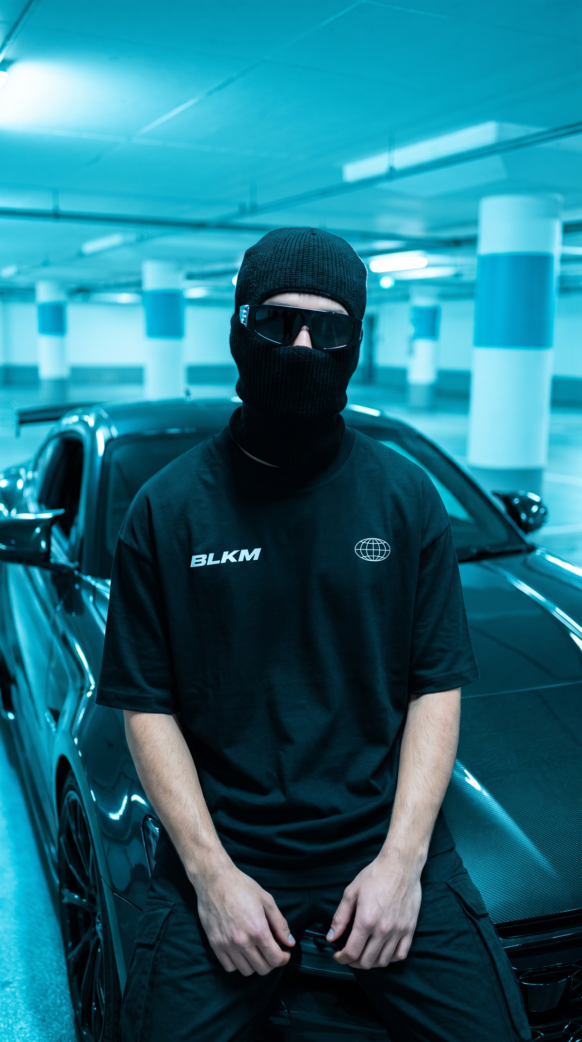 A person wearing a black balaclava, black sunglasses, and a black t-shirt with 'BLKM' printed on the chest sits in front of a black sports car inside a blue-lit parking garage.